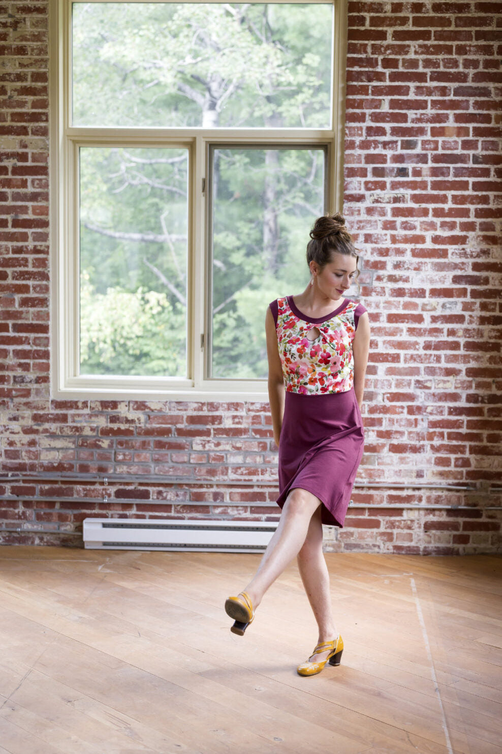 A woman wearing the Ocean Reversible Dress by Marie C in Cherry. The dress features a patterned bodice with a round neck and a cutout detail, cap sleeves, and a solid coloured skirt and opposite side bodice, with a fit and flare shape. She is standing in a room with a brick wall. 