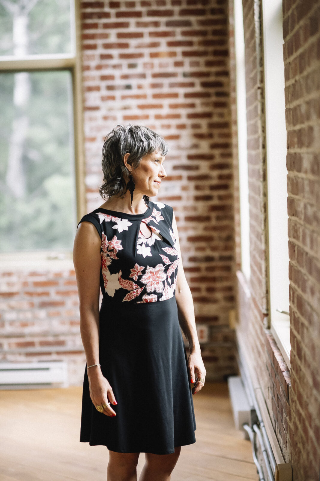 A woman wearing the Ocean Reversible Dress by Marie C in Black. The dress features a patterned bodice with a round neck and a cutout detail, cap sleeves, and a solid coloured skirt and opposite side bodice, with a fit and flare shape. She is standing in a room with a brick wall. 