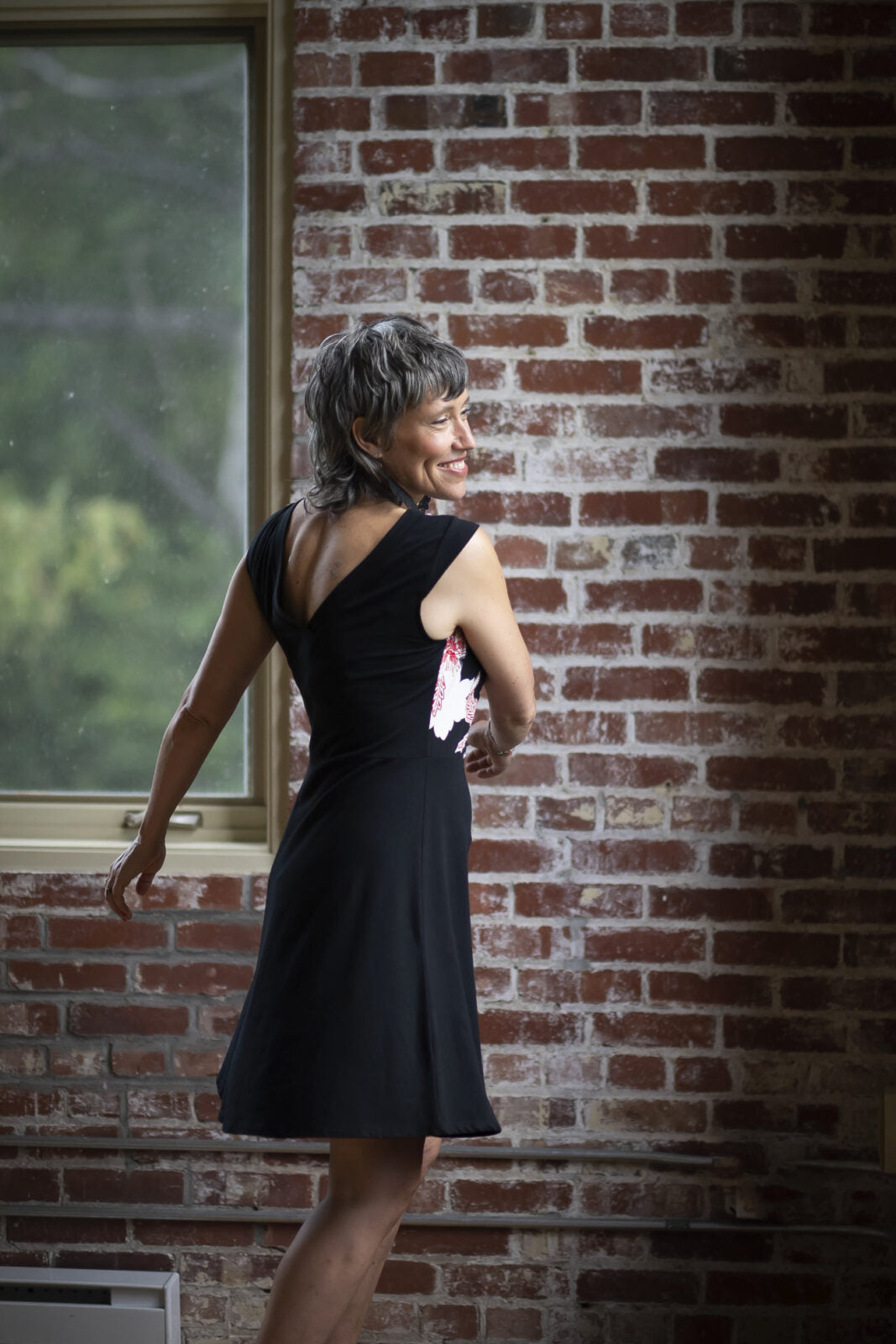 Side view of a woman wearing the Ocean Reversible Dress by Marie C in Black. The dress features a patterned bodice with a round neck and a cutout detail, cap sleeves, and a solid coloured skirt and opposite side bodice, with a fit and flare shape. She is standing in a room with a brick wall. 