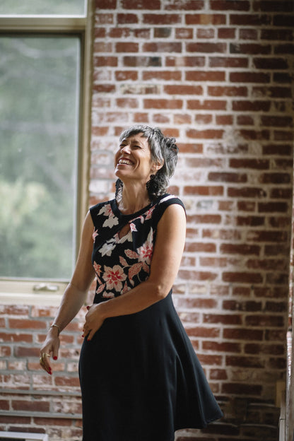 A woman wearing the Ocean Reversible Dress by Marie C in Black. The dress features a patterned bodice with a round neck and a cutout detail, cap sleeves, and a solid coloured skirt and opposite side bodice, with a fit and flare shape. She is standing in a room with a brick wall. 