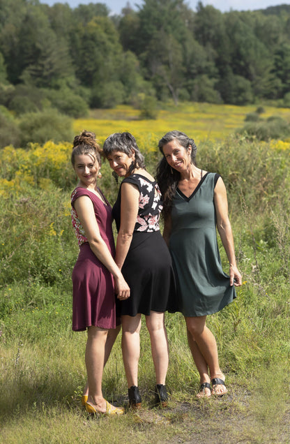 Three women wearing the Ocean Reversible Dress by Marie C. The dress features a patterned bodice with a round neck and a cutout detail, cap sleeves, and a solid coloured skirt and opposite side bodice, with a fit and flare shape. They are standing in a field.
