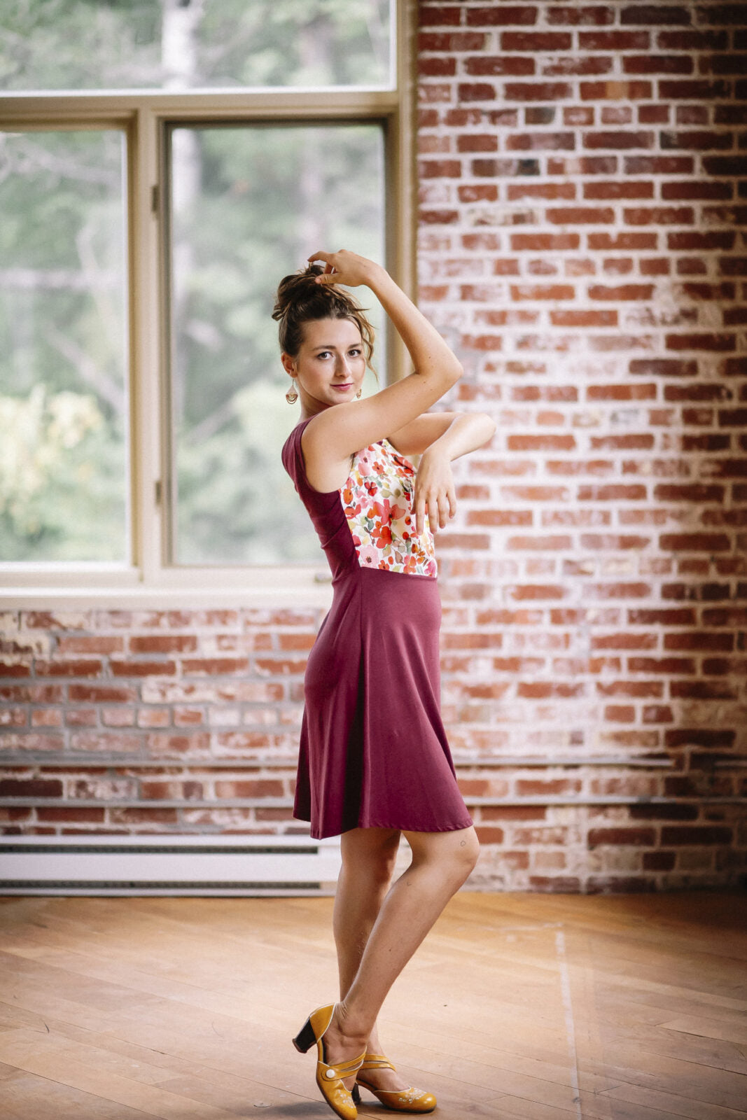 Side view of a woman wearing the Ocean Reversible Dress by Marie C in Cherry. The dress features a patterned bodice with a round neck and a cutout detail, cap sleeves, and a solid coloured skirt and opposite side bodice, with a fit and flare shape. She is standing in a room with a brick wall. 