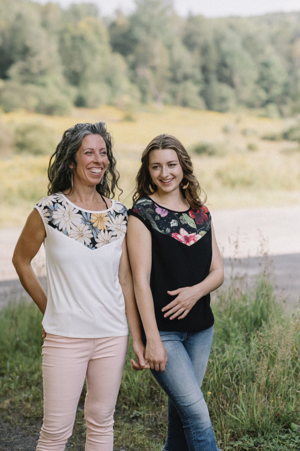 Two women wearing the Marie C Pepita Reversible Top, one in Ivory and one in Black Eyelet. The cap-sleeved top features patterned fabric across the chest on one side with solid colour on the other side. They are standing in a field. 