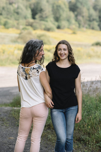 Two women wearing the Marie C Pepita Reversible Top, one in Ivory and one in Black Eyelet. The cap-sleeved top features patterned fabric across the chest on one side with solid colour on the other side. They are standing on a gravel road.
