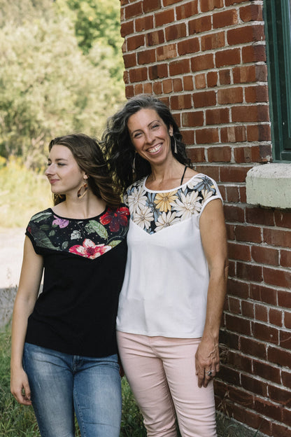 Two women wearing the Marie C Pepita Reversible Top, one in Ivory and one in Black Eyelet. The cap-sleeved top features patterned fabric across the chest on one side with solid colour on the other side. They are standing beside a brick building.
