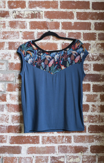 The Marie C Pepita Reversible Top in Blue is shown hanging against a brick wall. The cap-sleeved top features patterned fabric across the chest on one side with solid colour on the other side. 