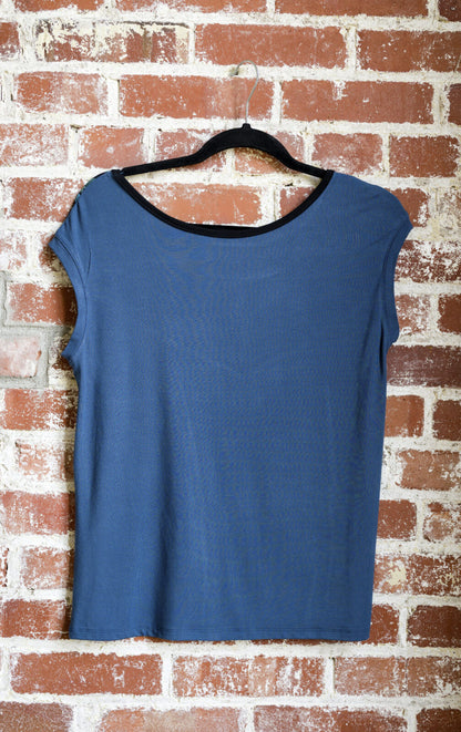 The Marie C Pepita Reversible Top in Blue is shown hanging against a brick wall. The cap-sleeved top features patterned fabric across the chest on one side with solid colour on the other side. 