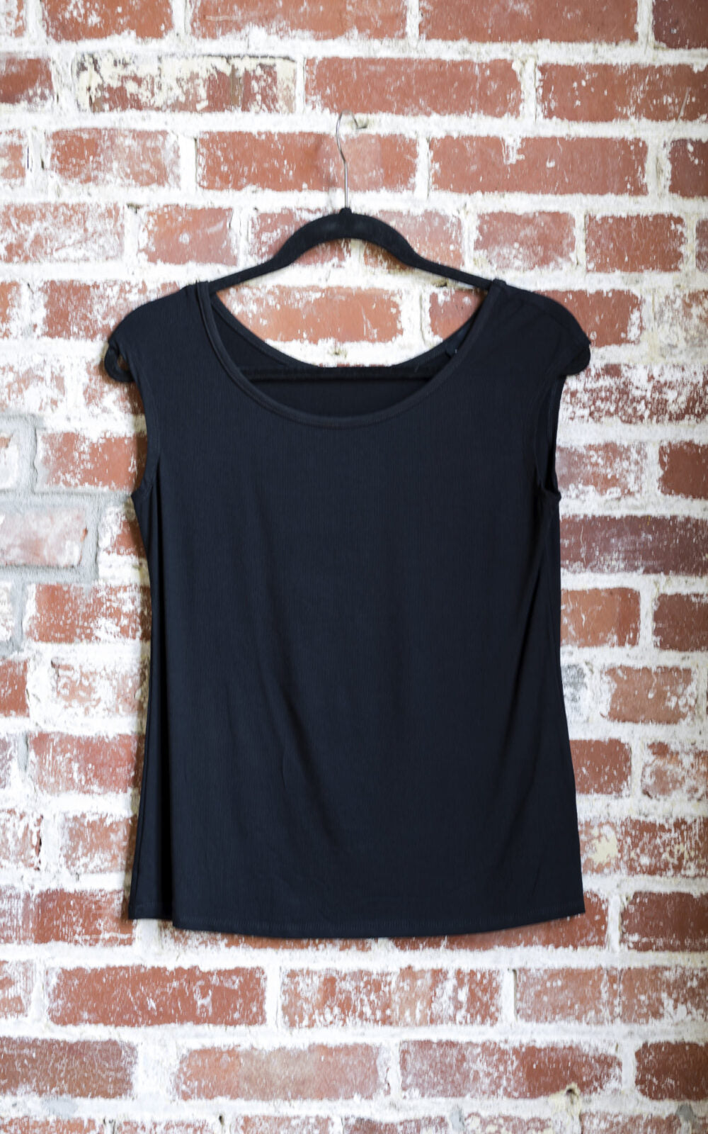 The Sol Top by Marie C in Black, with a scoop neck and cap sleeves, is shown hanging against a brick wall. 