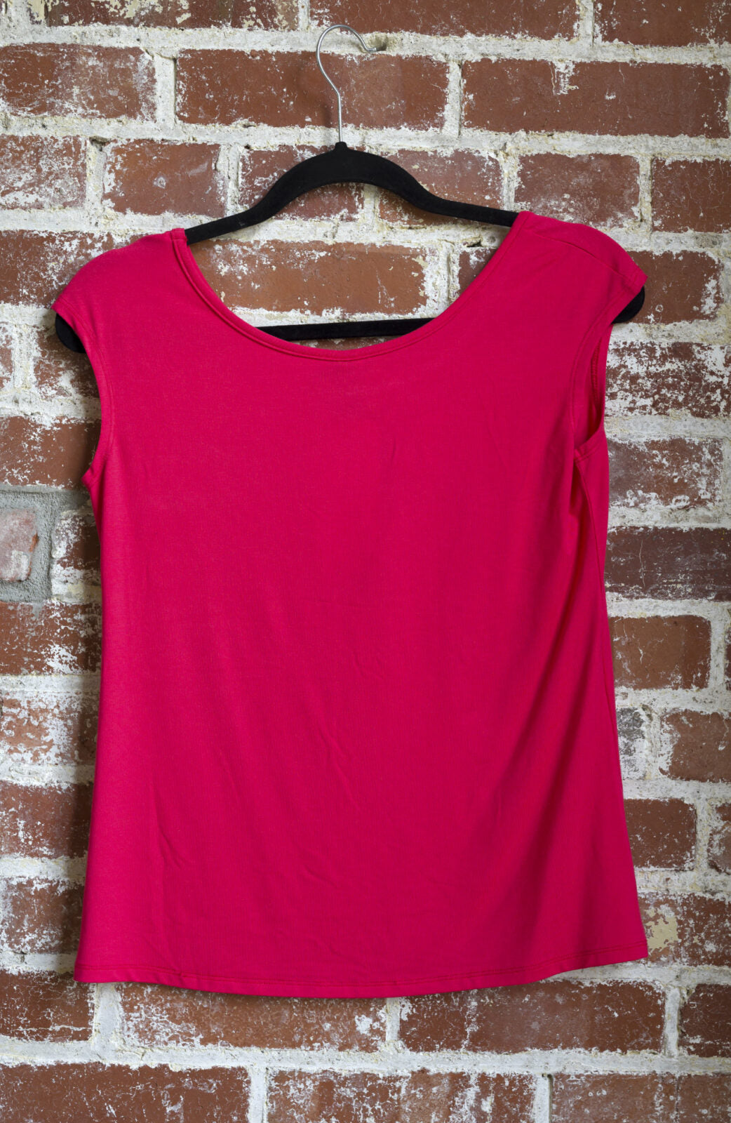 A back view of the Sol Top from Marie C in Coral, a reversible cap sleeve tee, is shown hanging vertically in front of a brick wall.