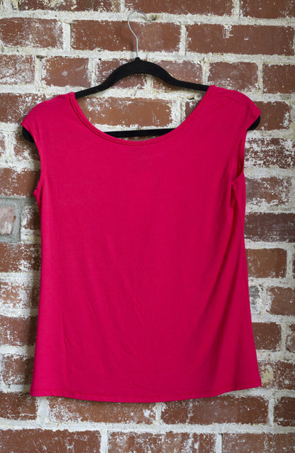 A back view of the Sol Top from Marie C in Coral, a reversible cap sleeve tee, is shown hanging vertically in front of a brick wall.