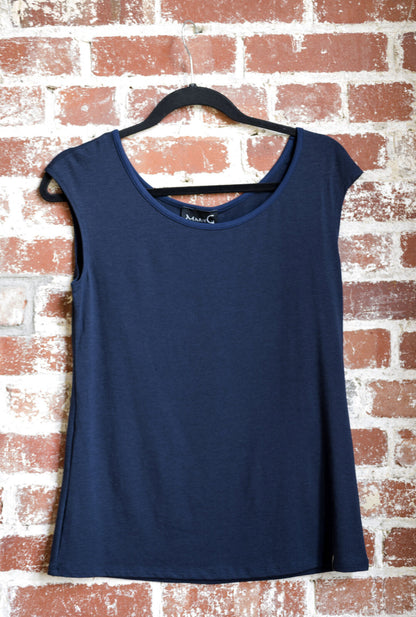 The Sol Top by Marie C in Navy, with a scoop neck and cap sleeves, is shown hanging against a brick wall. 