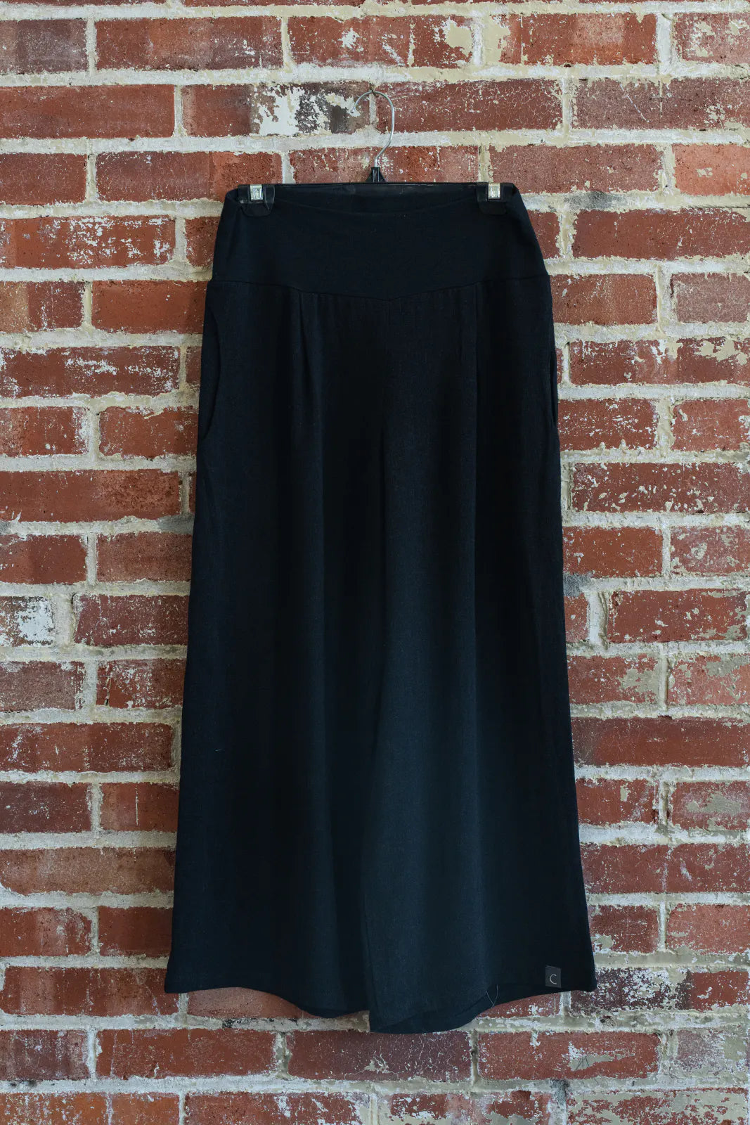 The Verano Pants by Marie C in Black are shown hanging in front of a brick wall. 
