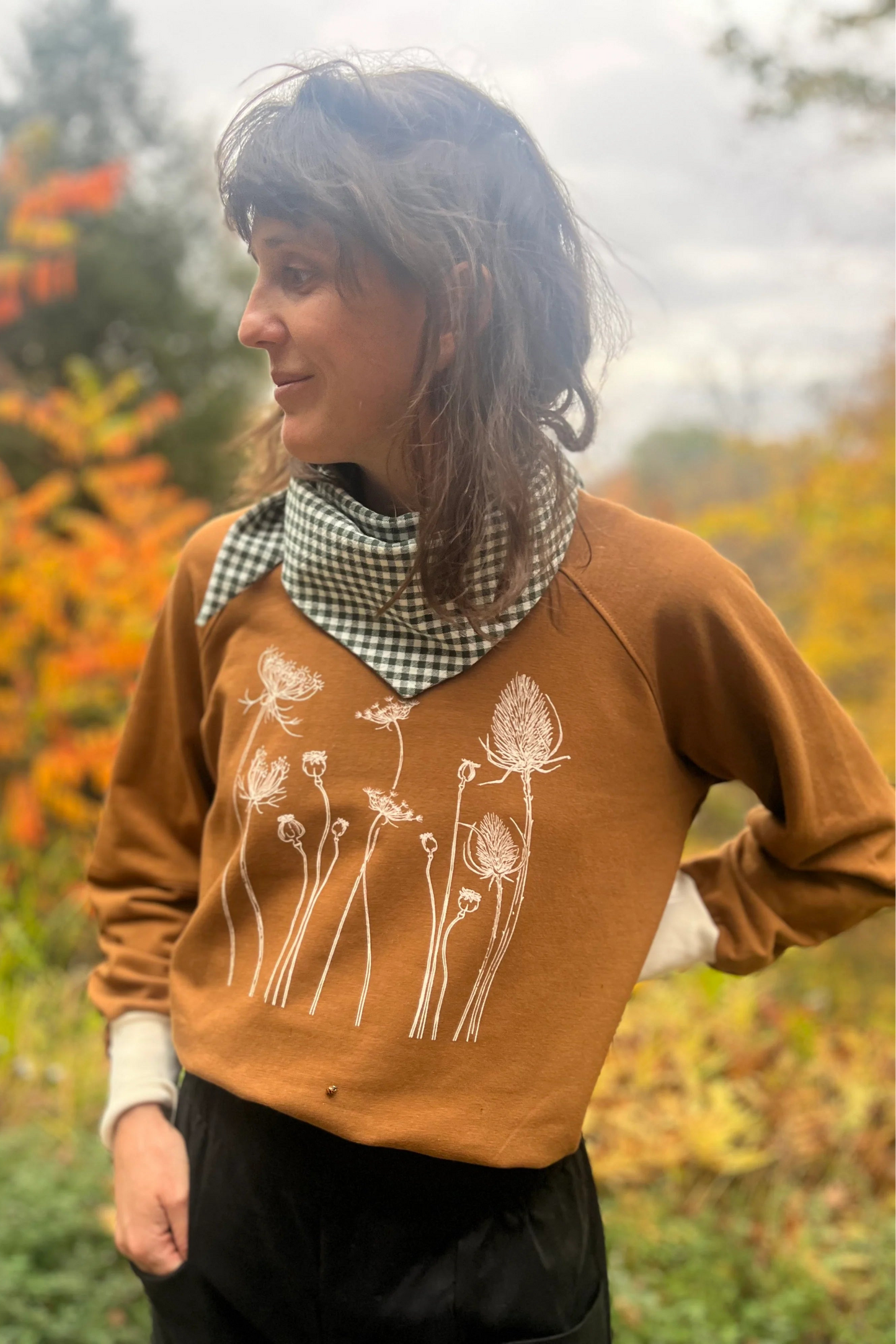 A woman wearing the Marilou Crewneck with Flowers by Kazak in Teak, a raglan sleeved sweatshirt with a screen print design. She is wearing it with jeans and standing in a garden. 
