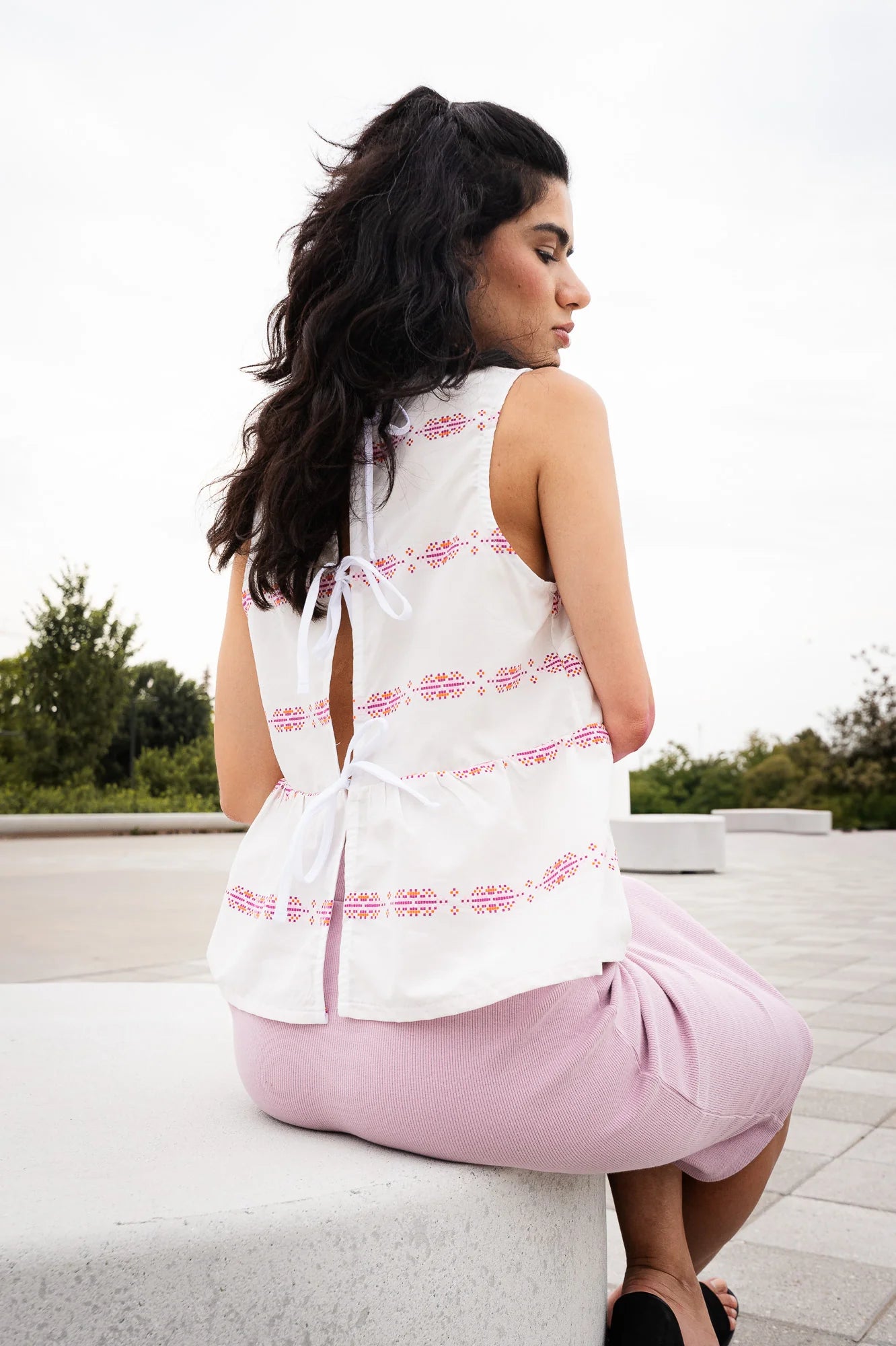 Back view of a woman wearing the Malé Top from MAS, a sleeveless top with a peplum and ties up the back. She is wearing it with wide-leg pants and sitting outdoors. 