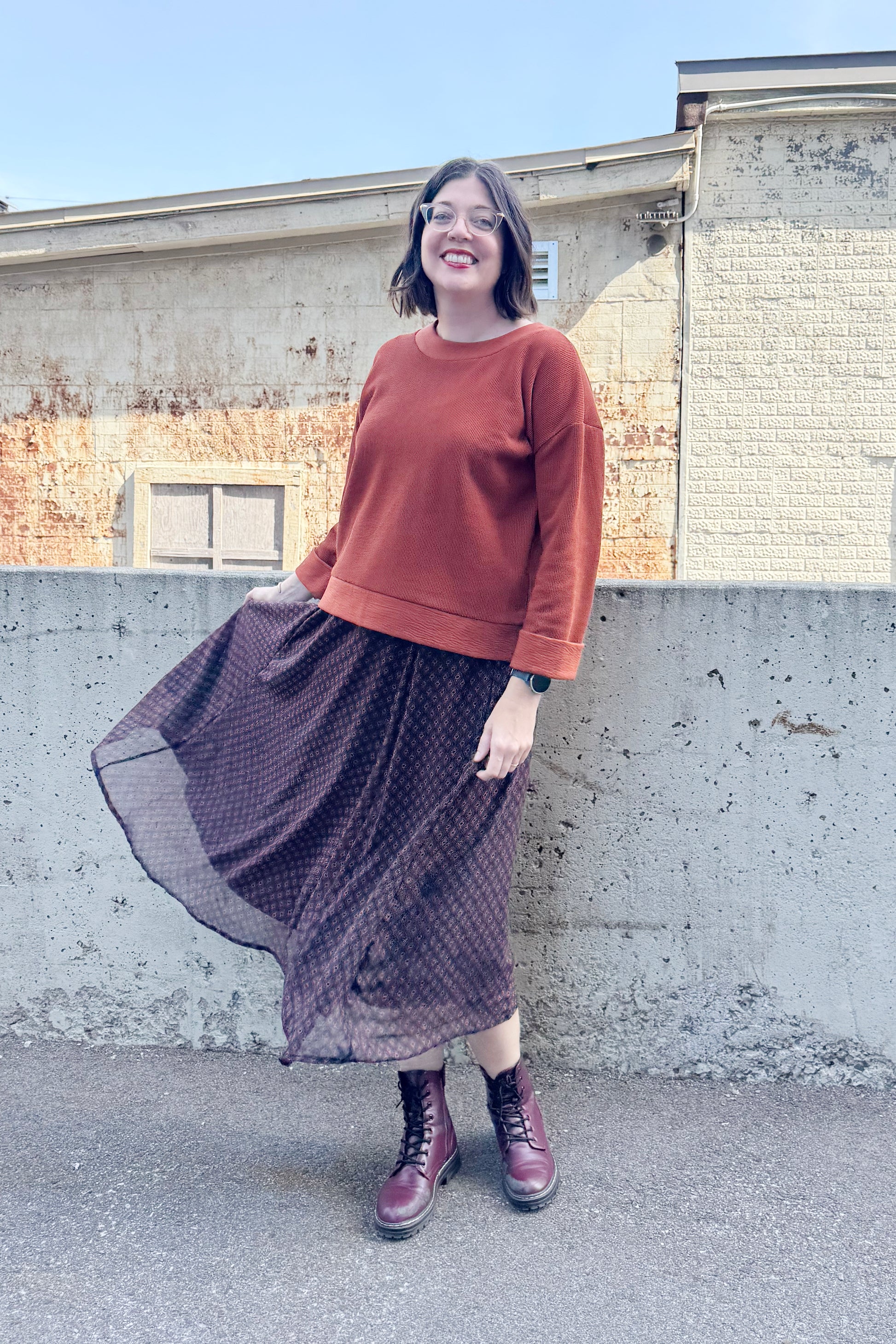 A woman wearing the Espresso Skirt by Cherry Bobin in Black and Brown print, a long and flowing chiffon skirt that's fully lined. She's wearing it with the Mocha Sweater in Bronze and standing outside in front of a brick building.
