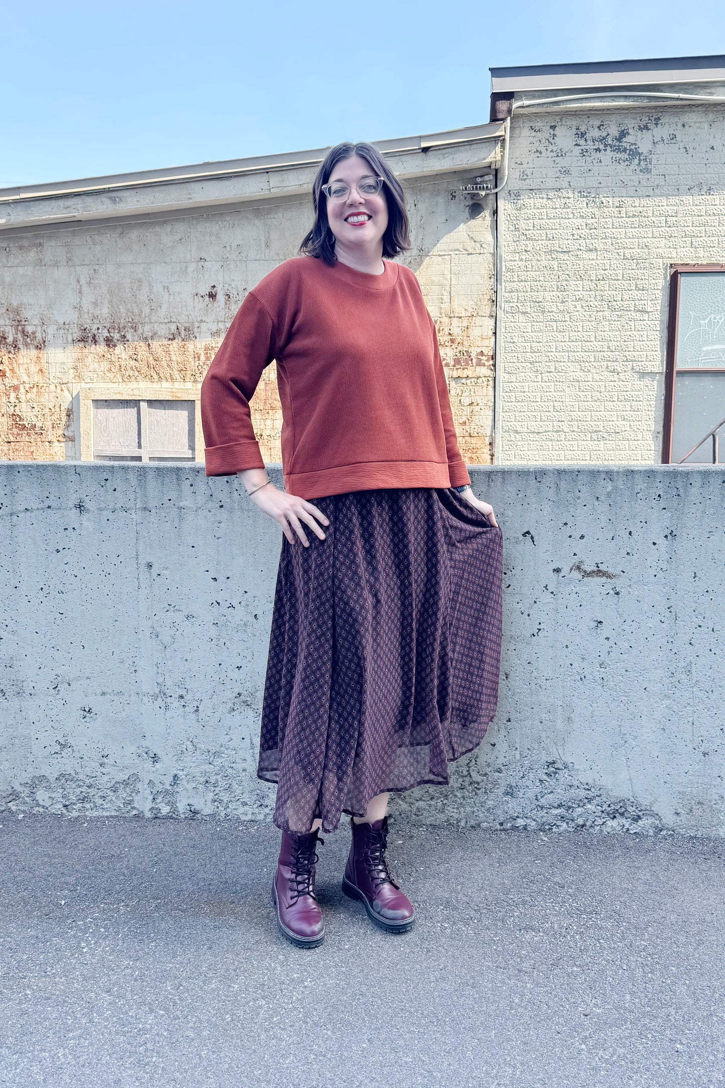 A woman wearing the Espresso Skirt by Cherry Bobin in Black and Brown print, a long and flowing chiffon skirt that's fully lined. She's wearing it with the Mocha Sweater in Bronze and standing outside in front of a brick building.
