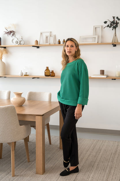 A woman wearing the Manhattan Pants by Cherry Bobin in Grey with the Mocha Sweater in Green and standing in a room with shelves. 