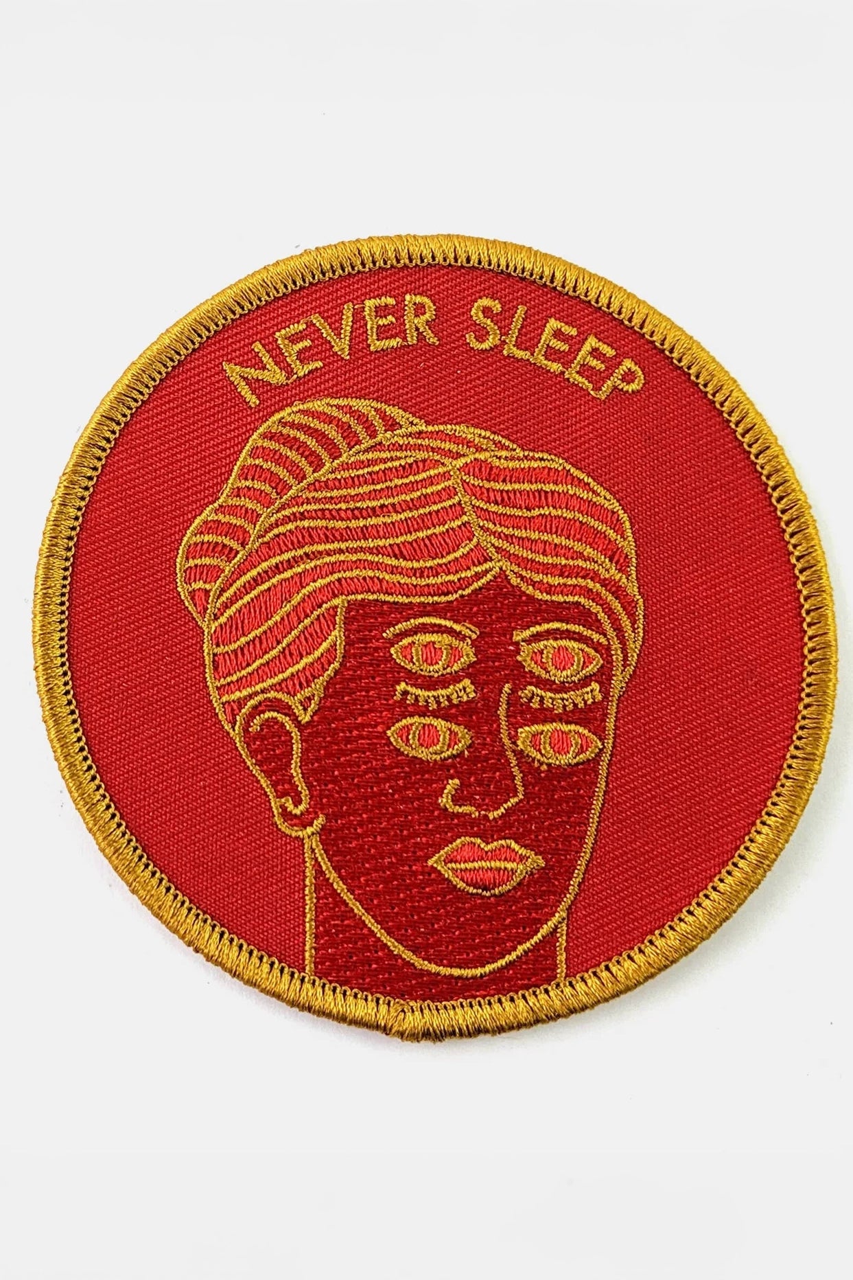 The Never Sleep Patch from Badaboöm Studio, circular embroidered patch with a red background, gold border, and a stylized face with two sets of open eyes. The words 'NEVER SLEEP' are embroidered in gold at the top.