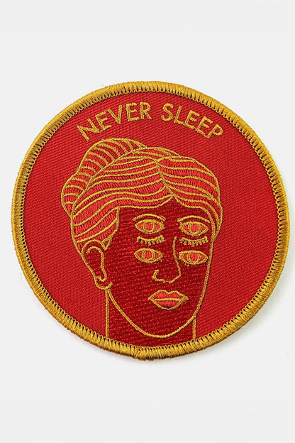 The Never Sleep Patch from Badaboöm Studio, circular embroidered patch with a red background, gold border, and a stylized face with two sets of open eyes. The words 'NEVER SLEEP' are embroidered in gold at the top.
