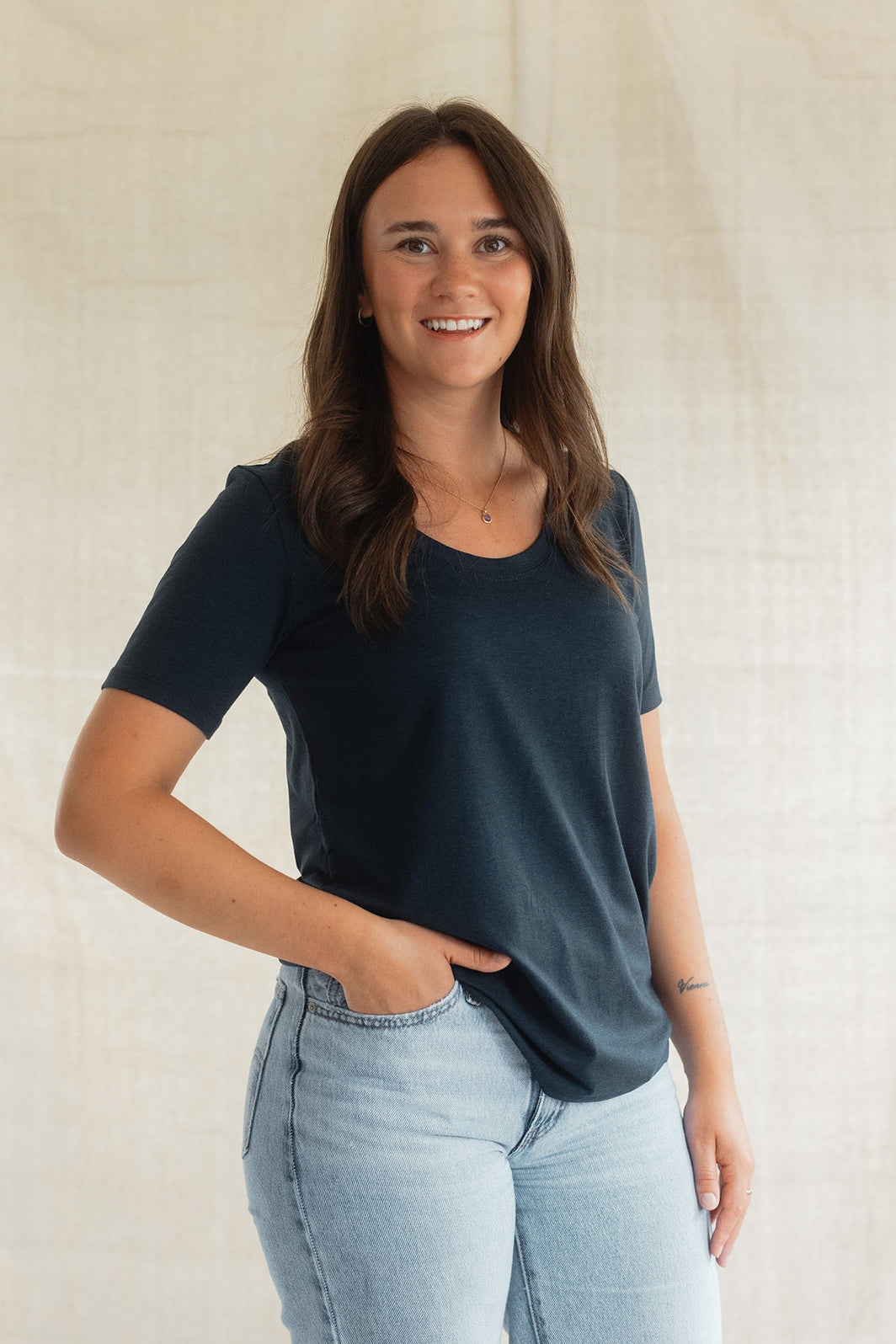 A woman wearing the Rideau Shoreline tee by Blondie in Navy, with a scoop neck and short sleeves. She is wearing it with jeans and standing in front of a beige background. 