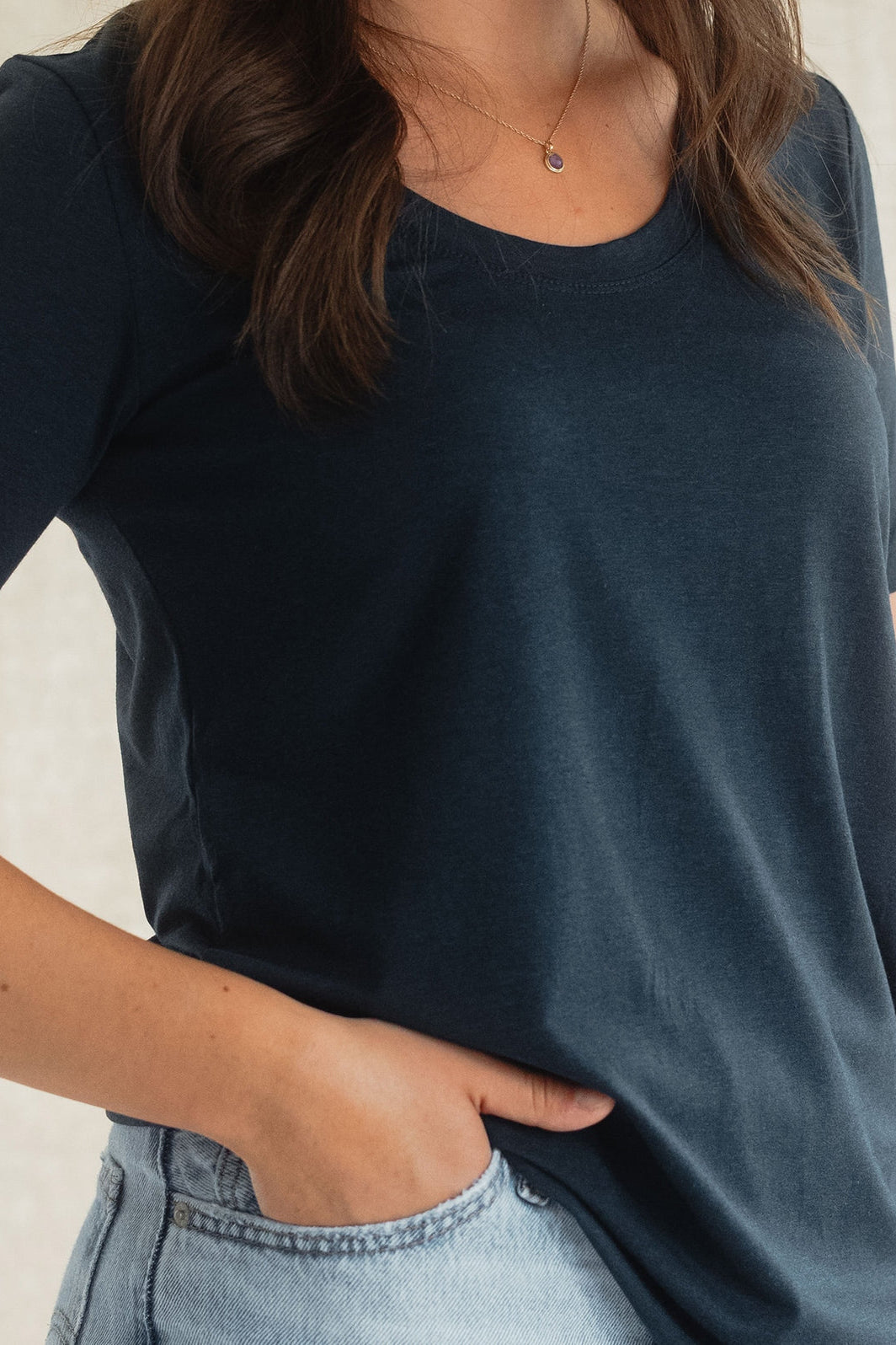 Close up of a woman wearing the Rideau Shoreline tee by Blondie in Navy, with a scoop neck and short sleeves. She is wearing it with jeans and standing in front of a beige background. 