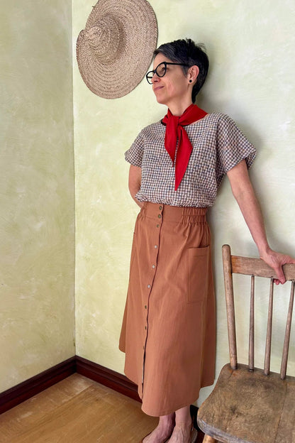 A woman wearing the Nymphea Skirt from Kazak in Ginger, a midi length A-line skirt with an elastic waist, brass buttons down the front, and large patch pockets. She is wearing it with a checkered top and standing in a room with a wooden chair.