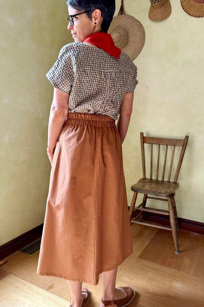 Back view of a woman wearing the Nymphea Skirt from Kazak in Ginger, a midi length A-line skirt with an elastic waist, brass buttons down the front, and large patch pockets. She is wearing it with a checkered top and standing in a room with a wooden chair.