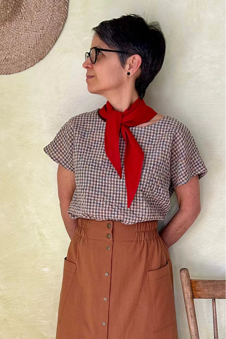 A woman wearing the Nymphea Skirt from Kazak in Ginger, a midi length A-line skirt with an elastic waist, brass buttons down the front, and large patch pockets. She is wearing it with a checkered top and standing in a room with a wooden chair.
