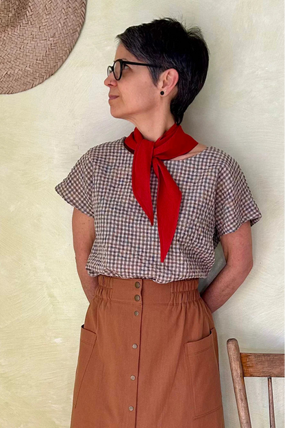 A woman wearing the Nymphea Skirt from Kazak in Ginger, a midi length A-line skirt with an elastic waist, brass buttons down the front, and large patch pockets. She is wearing it with a checkered top and standing in a room with a wooden chair.