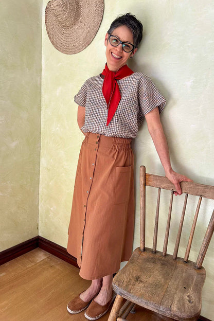 A woman wearing the Nymphea Skirt from Kazak in Ginger, a midi length A-line skirt with an elastic waist, brass buttons down the front, and large patch pockets. She is wearing it with a checkered top and standing in a room with a wooden chair.