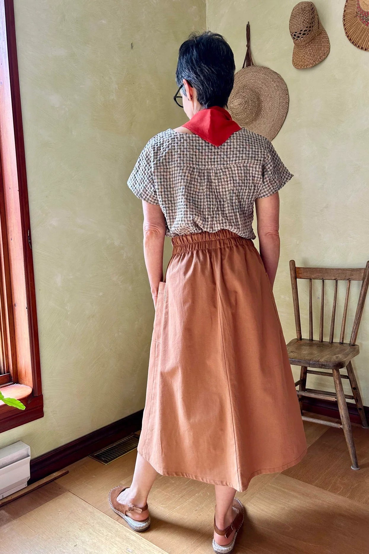 Back view of a woman wearing the Nymphea Skirt from Kazak in Ginger, a midi length A-line skirt with an elastic waist, brass buttons down the front, and large patch pockets. She is wearing it with a checkered top and standing in a room with a wooden chair.