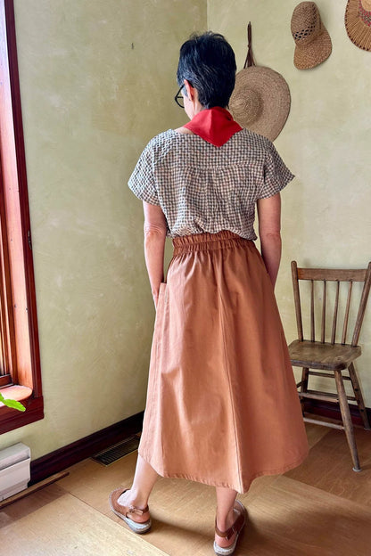 Back view of a woman wearing the Nymphea Skirt from Kazak in Ginger, a midi length A-line skirt with an elastic waist, brass buttons down the front, and large patch pockets. She is wearing it with a checkered top and standing in a room with a wooden chair.
