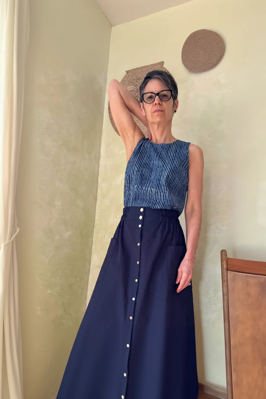 A woman wearing the Nymphea Skirt from Kazak in Navy, a midi length A-line skirt with an elastic waist, brass buttons down the front, and large patch pockets. She is wearing it with the Forsythia Top in Indigo stripe and standing in a room with a wooden chair.