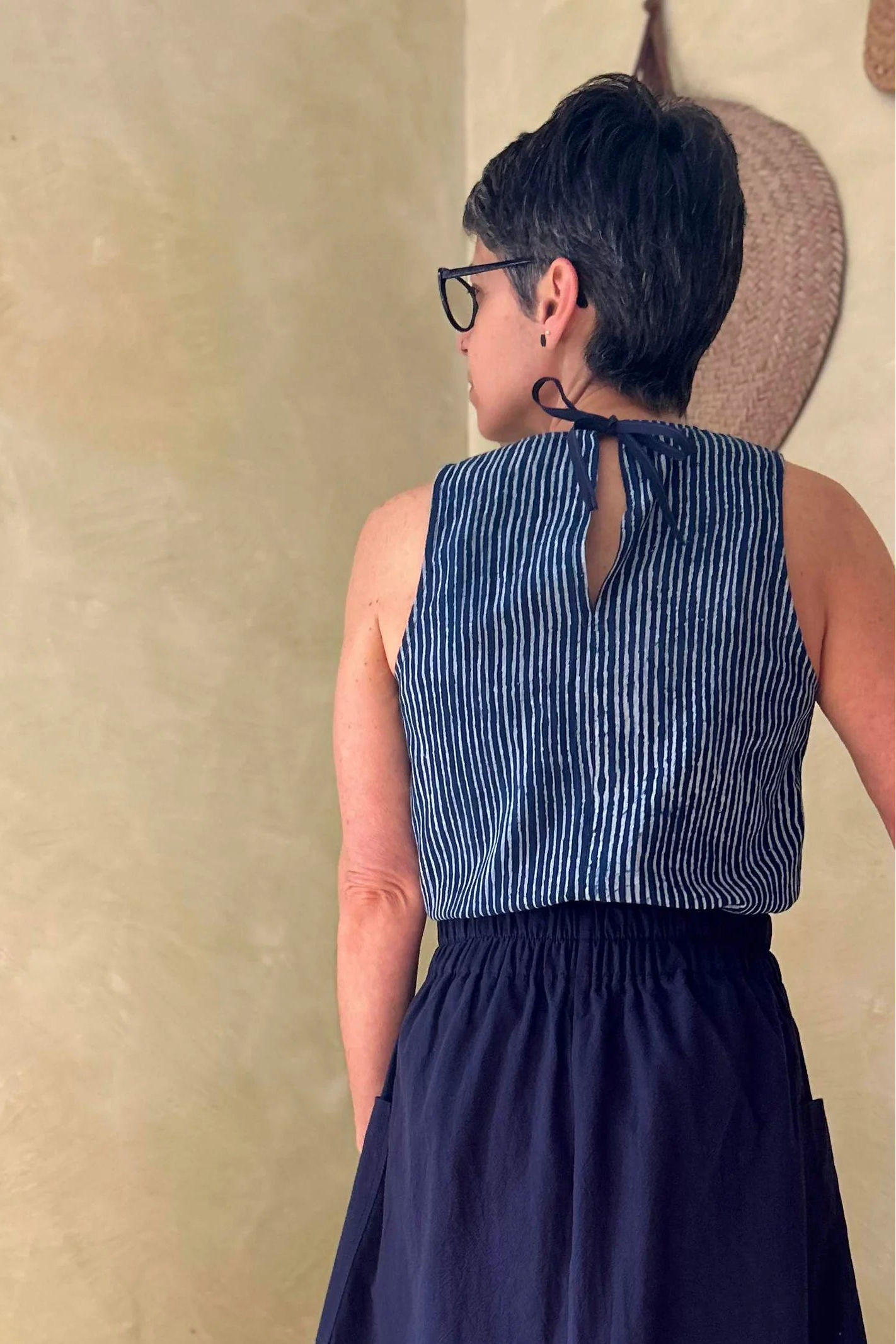 Back view of a woman wearing the Nymphia Skirt from Kazka in Navy, a midi length A-line skirt with elastic at the side waist, brass buttons down the front, and large patch pockets. She is wearing it with the Forsythia Top in Indigo stripe and standing in a room with a wooden chair.