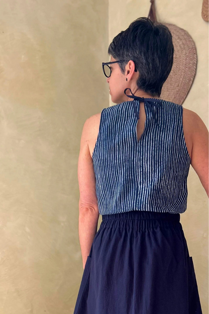 Back view of a woman wearing the Nymphia Skirt from Kazka in Navy, a midi length A-line skirt with elastic at the side waist, brass buttons down the front, and large patch pockets. She is wearing it with the Forsythia Top in Indigo stripe and standing in a room with a wooden chair.