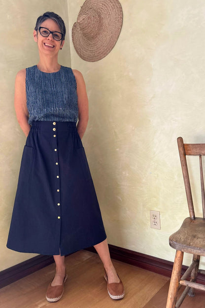 A woman wearing the Nymphea Skirt from Kazak in Navy, a midi length A-line skirt with an elastic waist, brass buttons down the front, and large patch pockets. She is wearing it with the Forsythia Top in Indigo stripe and standing in a room with a wooden chair. 