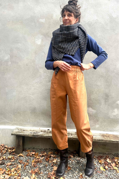 A woman wearing the Nyssa Pants by Kazak in Terracotta, barrel-legged pants with an elastic waistband and patch pockets. She is wearing them with the Oris Turtleneck in Indigo Waffle and standing outside by a grey wall. 