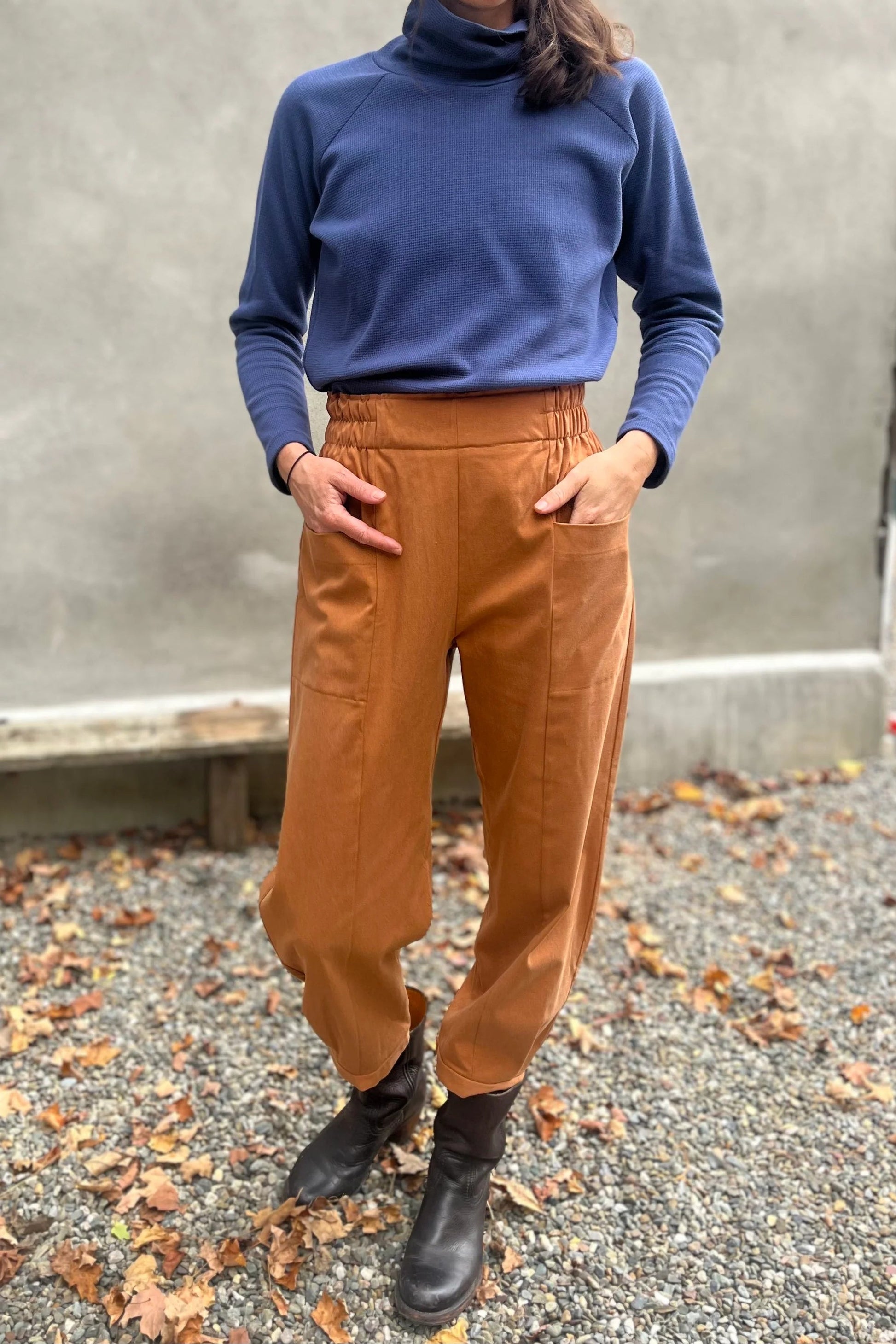 A woman wearing the Nyssa Pants by Kazak in Terracotta, barrel-legged pants with an elastic waistband and patch pockets. She is wearing them with the Oris Turtleneck in Indigo Waffle and standing outside by a grey wall. 
