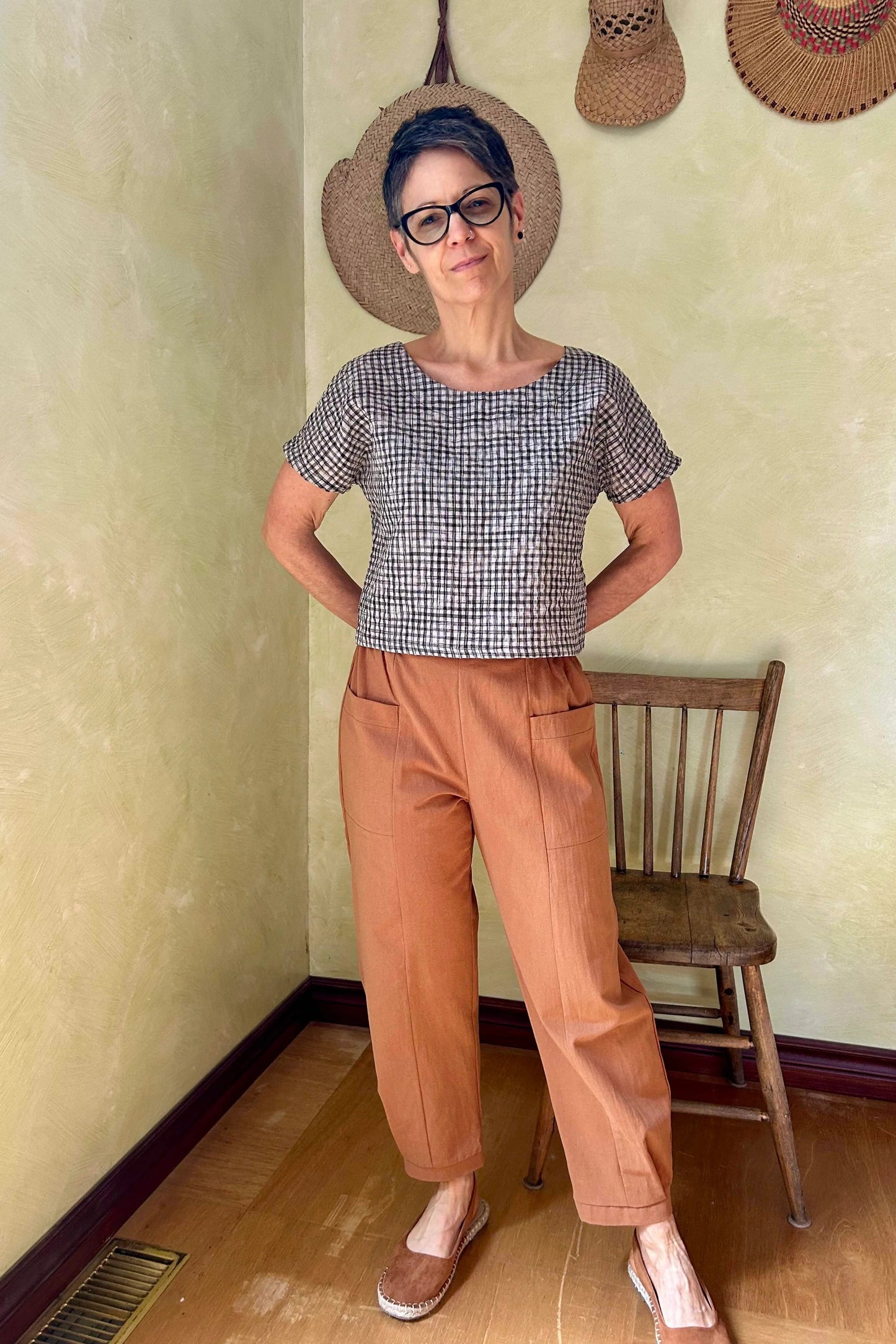 A woman wearing the Oxalis Pants from Kazak in Ginger, with a flat front waistband, legs that taper at the ankle, and large patch pockets. She is wearing them with a checkered top and standing in a room with a wooden chair. 