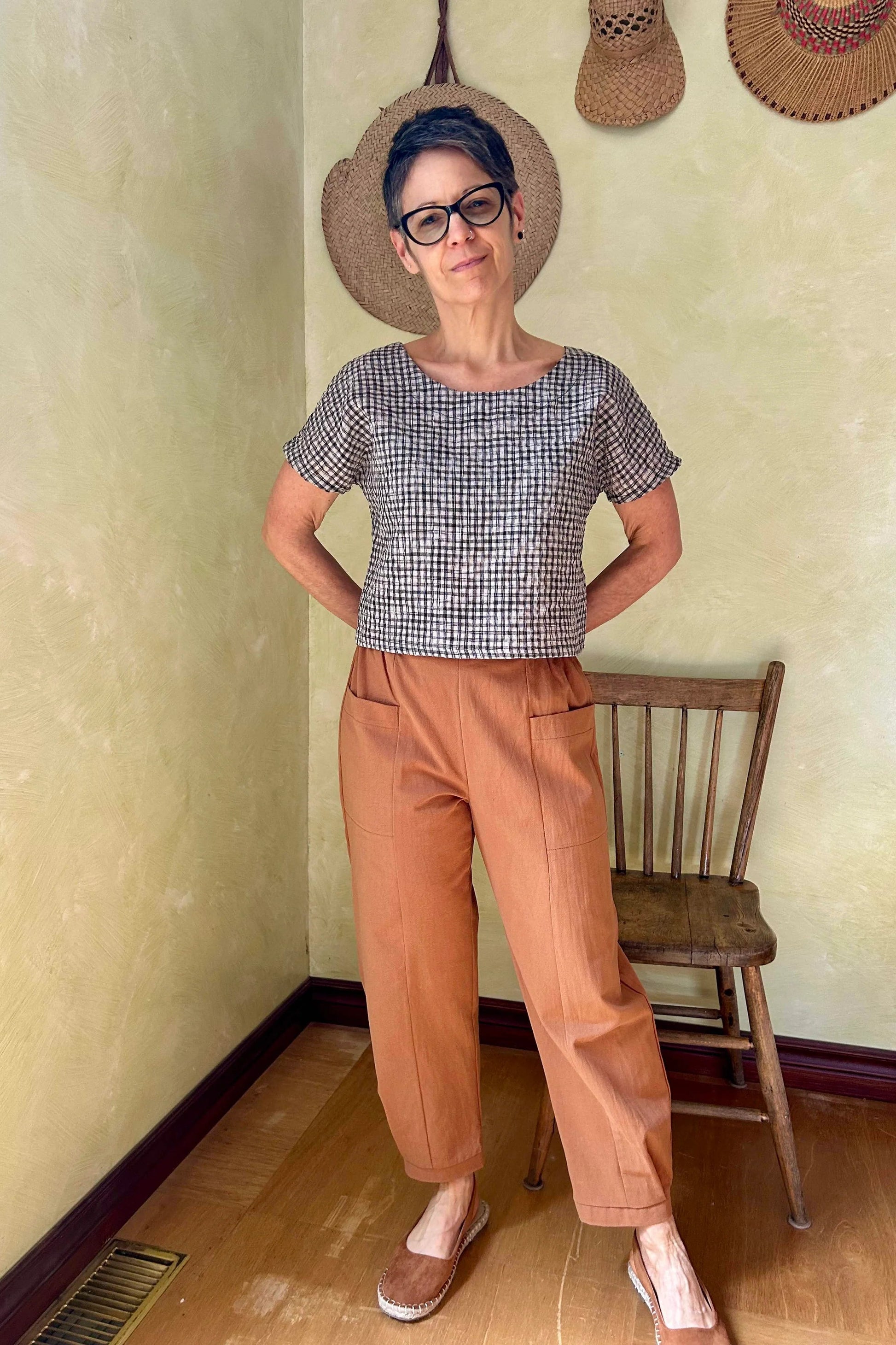 A woman wearing the Oxalis Pants from Kazak in Ginger, with a flat front waistband, legs that taper at the ankle, and large patch pockets. She is wearing them with a checkered top and standing in a room with a wooden chair. 
