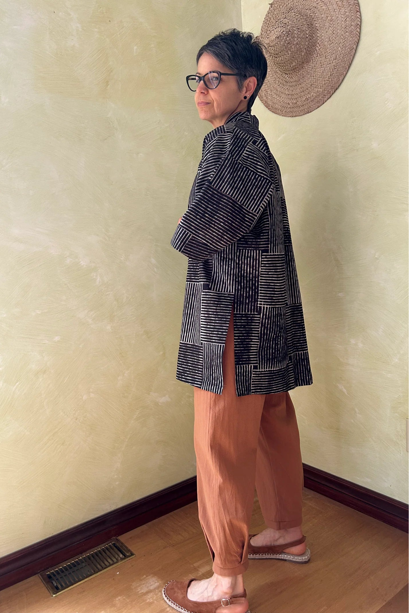 Side view of a woman wearing the Mirano Jacket by Kazak in Oris Slate, a cross hatch pattern. It is a  mid-thigh length open kimono jacket with 3/4 sleeves. She is wearing it with the Oxalis pants in Ginger and standing in a room by a wooden chair. 