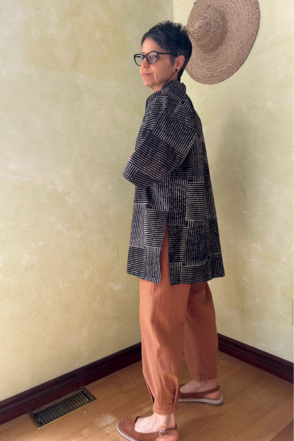 Side view of a woman wearing the Mirano Jacket by Kazak in Oris Slate, a cross hatch pattern. It is a  mid-thigh length open kimono jacket with 3/4 sleeves. She is wearing it with the Oxalis pants in Ginger and standing in a room by a wooden chair. 