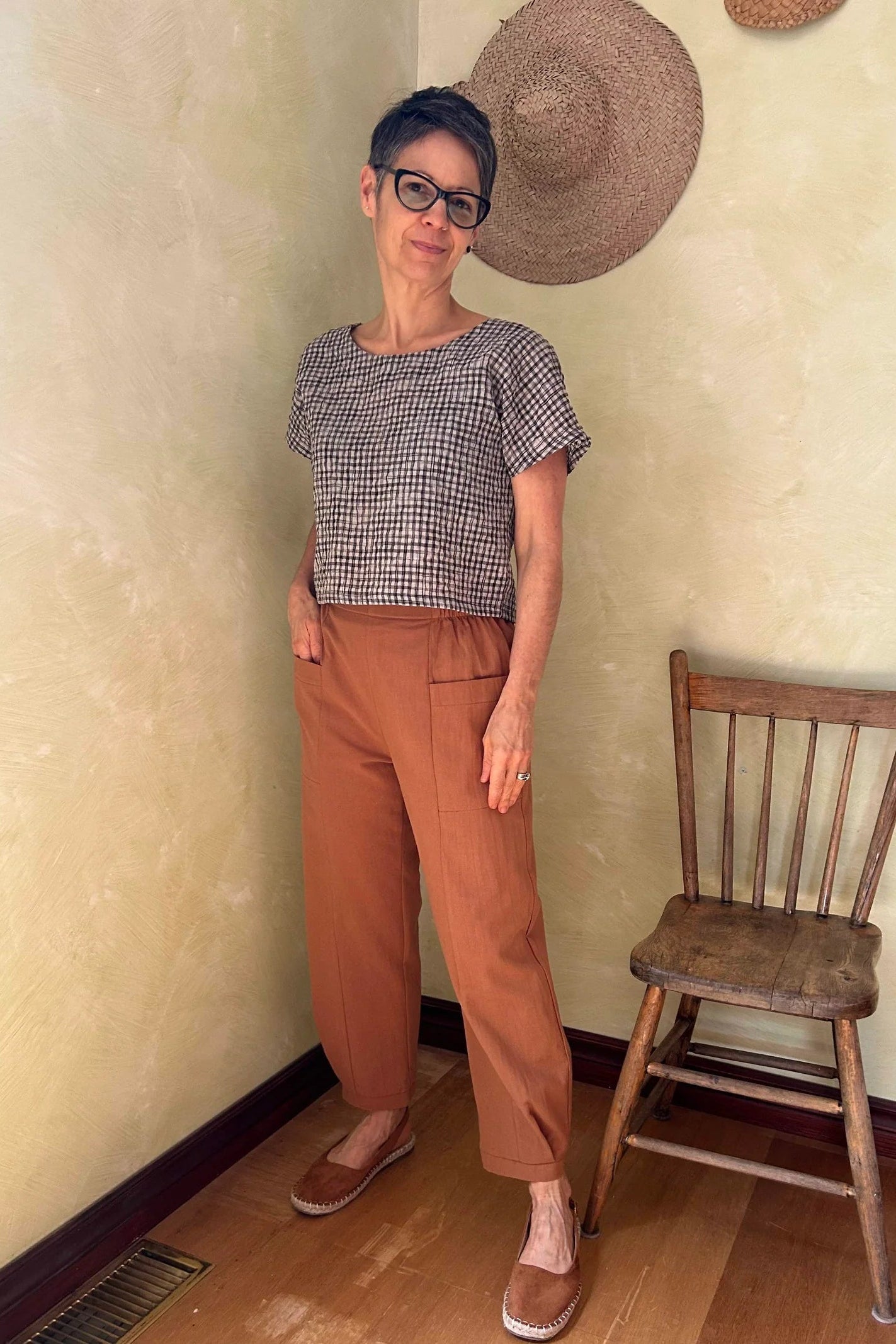 A woman wearing the Oxalis Pants from Kazak in Ginger, with a flat front waistband, legs that taper at the ankle, and large patch pockets. She is wearing them with a checkered top and standing in a room with a wooden chair.