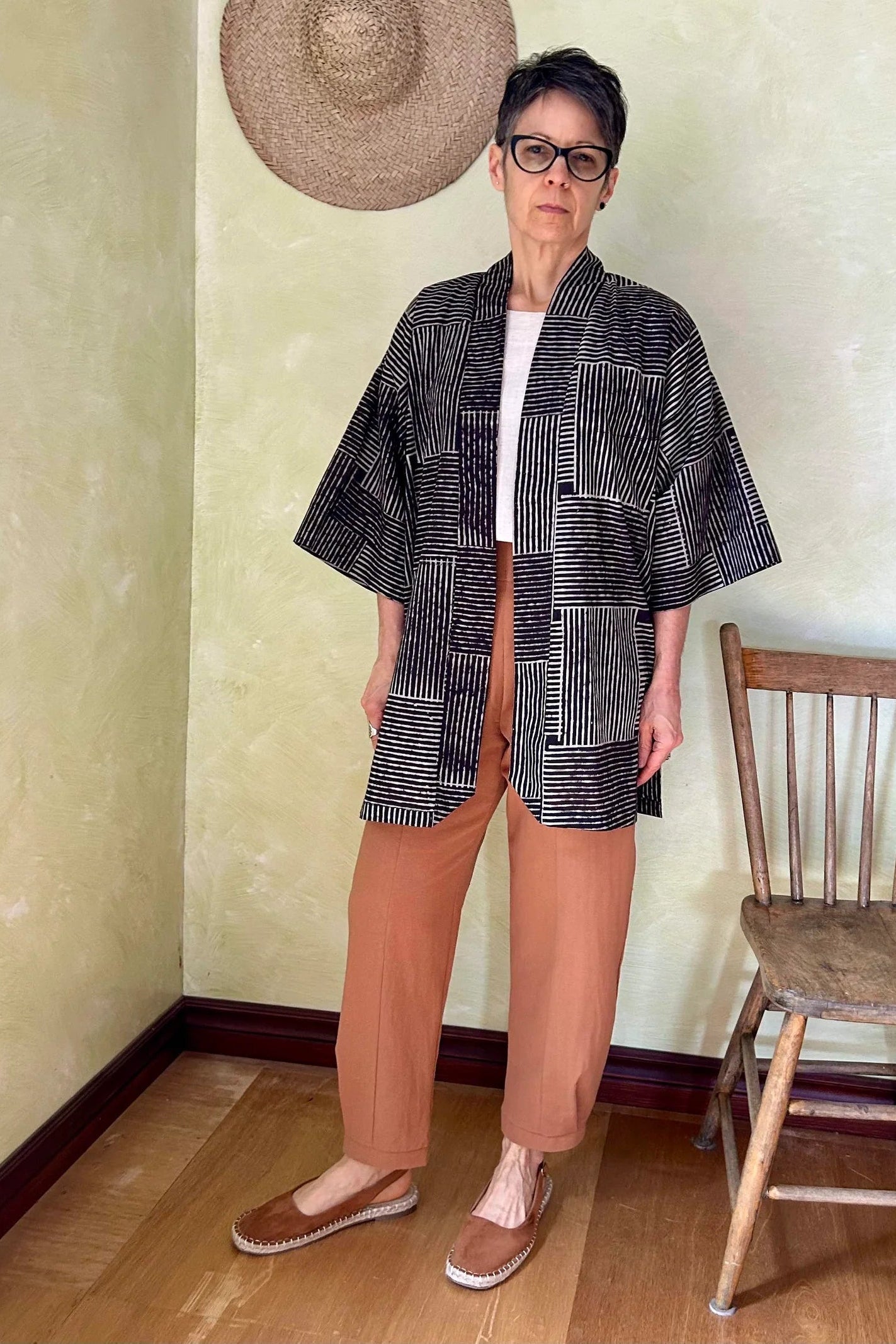 A woman wearing the Oxalis Pants from Kazak in Ginger, with a flat front waistband, legs that taper at the ankle, and large patch pockets. She is wearing them with a kimono style jacket and standing in a room with a wooden chair.