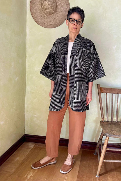 A woman wearing the Oxalis Pants from Kazak in Ginger, with a flat front waistband, legs that taper at the ankle, and large patch pockets. She is wearing them with a kimono style jacket and standing in a room with a wooden chair.