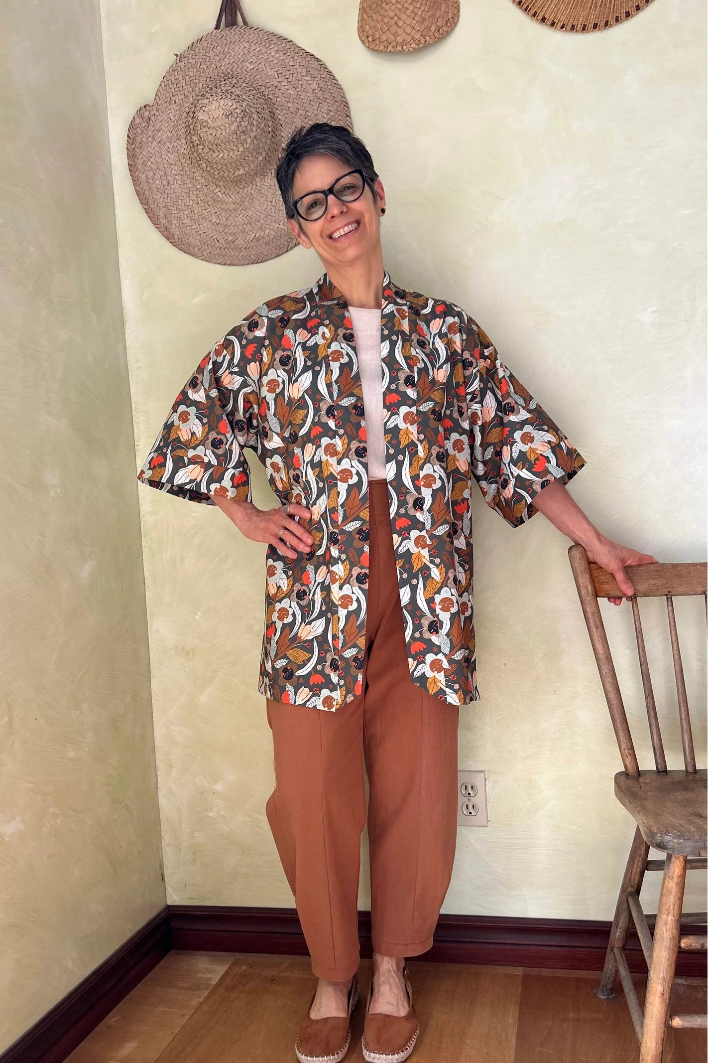 A woman wearing the Oxalis Pants from Kazak in Ginger, with a flat front waistband, legs that taper at the ankle, and large patch pockets. She is wearing them with a kimono style jacket and standing in a room with a wooden chair.

