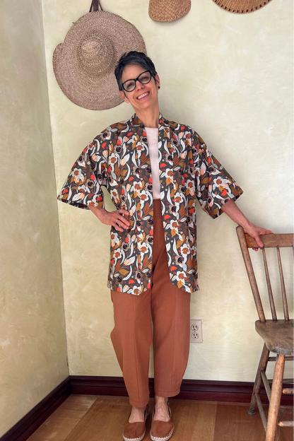 A woman wearing the Oxalis Pants from Kazak in Ginger, with a flat front waistband, legs that taper at the ankle, and large patch pockets. She is wearing them with a kimono style jacket and standing in a room with a wooden chair.

