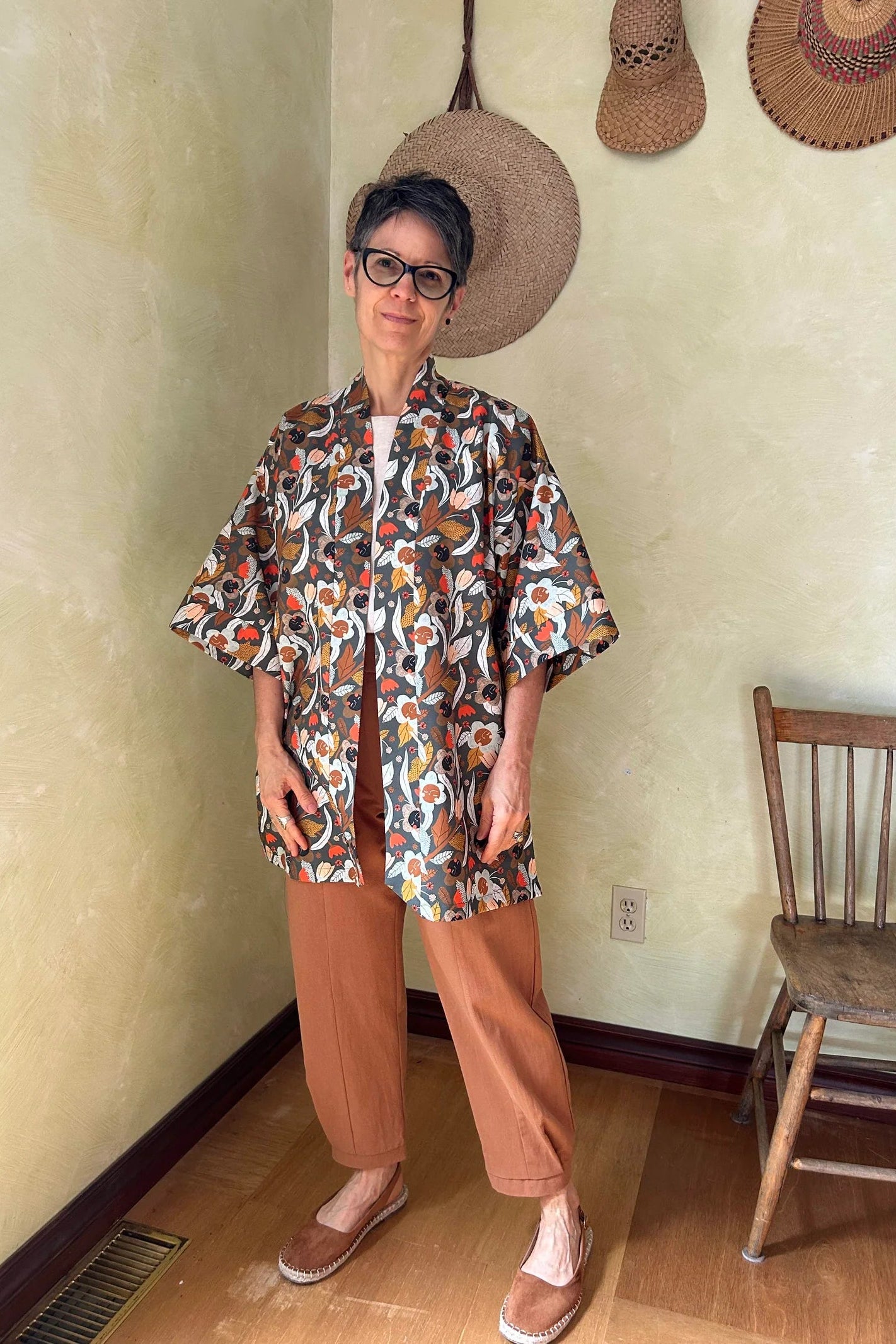 A woman wearing the Oxalis Pants from Kazak in Ginger, with a flat front waistband, legs that taper at the ankle, and large patch pockets. She is wearing them with a kimono style jacket and standing in a room with a wooden chair.