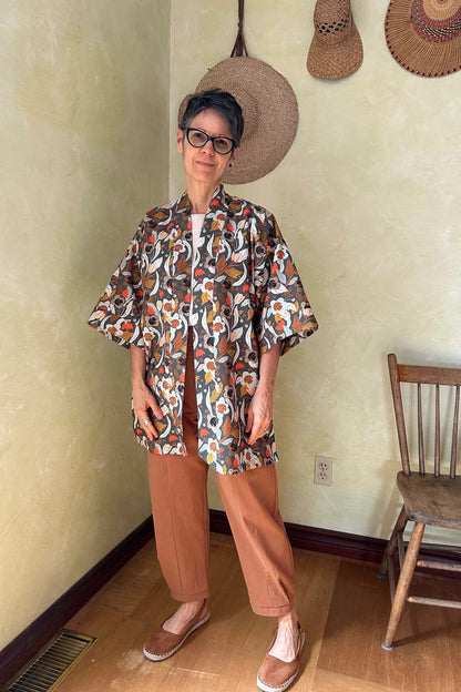 A woman wearing the Oxalis Pants from Kazak in Ginger, with a flat front waistband, legs that taper at the ankle, and large patch pockets. She is wearing them with a kimono style jacket and standing in a room with a wooden chair.