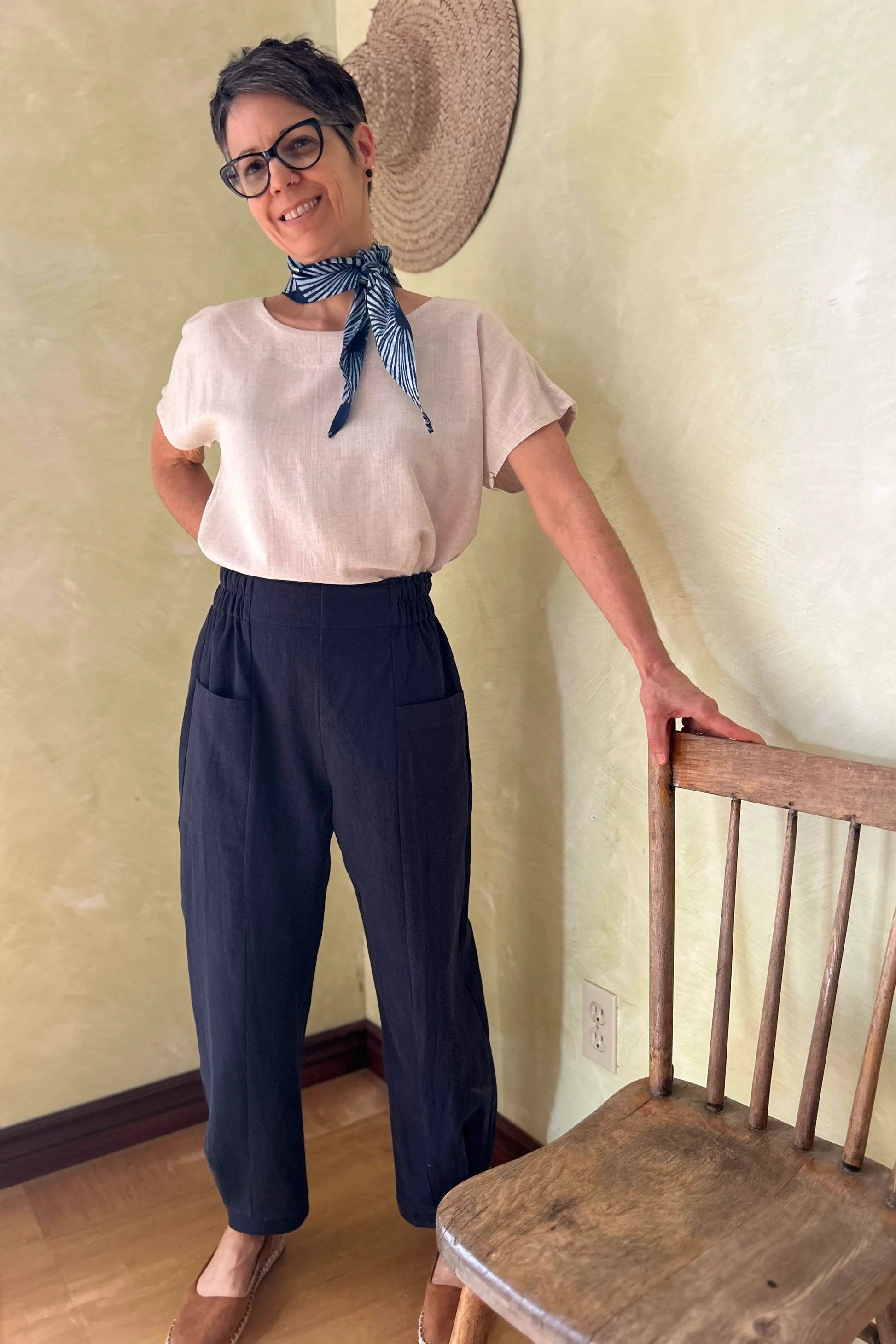 A woman wearing the Oxalis Pants from Kazak in Indigo, with a flat front waistband, legs that taper at the ankle, and large patch pockets. She is wearing them with a cream top and standing in a room with a wooden chair. 
